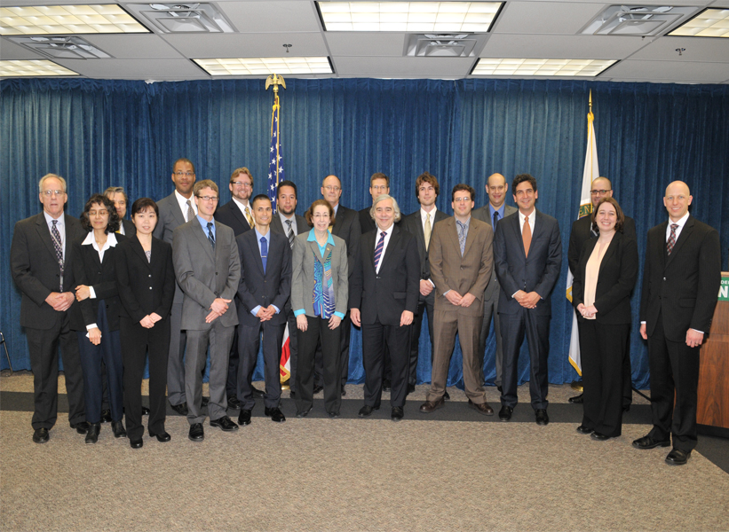 A group of 19 adults in suits, standing in front of conference drapes.